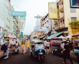 A bustling street filled with tuk-tuks and pedestrians, displaying colorful signage and vendors on either side. Buildings with multiple stories are packed closely along the street. The environment appears lively with a mix of locals and tourists, and the atmosphere is vibrant with a sense of daily life activity.