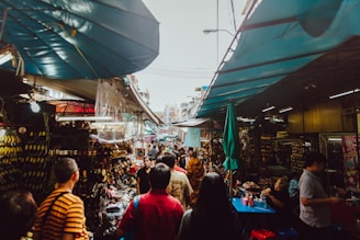 A bustling outdoor market with stalls lining both sides, covered by tarps. Various goods like shoes and accessories are displayed. People are walking and shopping, creating a lively atmosphere with a mix of colors and activities.
