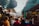 A bustling outdoor market with stalls lining both sides, covered by tarps. Various goods like shoes and accessories are displayed. People are walking and shopping, creating a lively atmosphere with a mix of colors and activities.