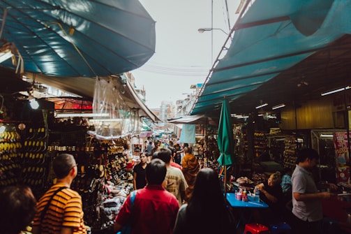 A bustling outdoor market with stalls lining both sides, covered by tarps. Various goods like shoes and accessories are displayed. People are walking and shopping, creating a lively atmosphere with a mix of colors and activities.