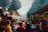 A bustling outdoor market with stalls lining both sides, covered by tarps. Various goods like shoes and accessories are displayed. People are walking and shopping, creating a lively atmosphere with a mix of colors and activities.