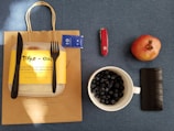 A brown paper bag with a take-out container, black knife, and fork on top sits on a blue surface. To the right are a pack of condiments, a red Swiss Army knife, a whole pomegranate, a mug filled with blueberries, and a smartphone.