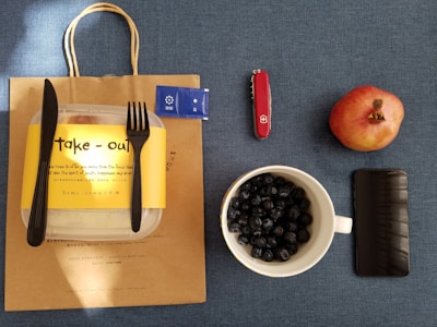 A brown paper bag with a take-out container, black knife, and fork on top sits on a blue surface. To the right are a pack of condiments, a red Swiss Army knife, a whole pomegranate, a mug filled with blueberries, and a smartphone.