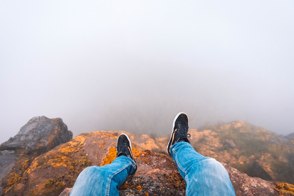 person wearing blue denim jeans sitting on brown wooden rock looking down on mist