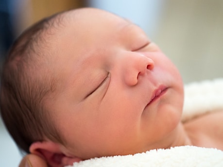 A newborn baby is peacefully sleeping, wrapped in a soft, cream-colored blanket. The close-up captures its serene expression with closed eyes and soft features, highlighting its gentle skin and faint hairline.
