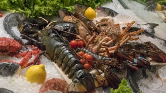 A display of fresh seafood arranged on a bed of ice. Large lobsters with rubber bands on their claws are surrounded by fish, crabs, and shellfish. Red cherry tomatoes and yellow lemons add color to the composition, while green leafy lettuce provides a vibrant backdrop.