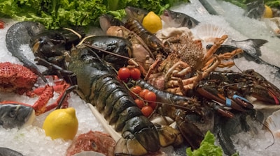 A display of fresh seafood arranged on a bed of ice. Large lobsters with rubber bands on their claws are surrounded by fish, crabs, and shellfish. Red cherry tomatoes and yellow lemons add color to the composition, while green leafy lettuce provides a vibrant backdrop.