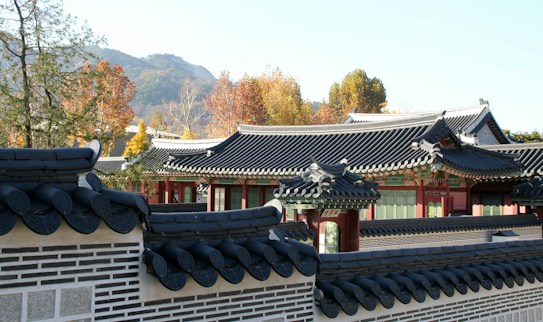 Traditional Korean architecture featuring intricately designed rooftops with dark tiles and ornate details. The setting includes colorful autumn foliage and a background of distant mountains under a clear sky.
