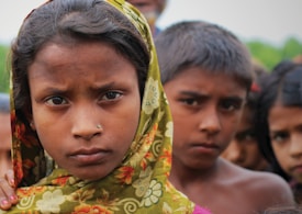 A group of children is portrayed with one girl prominently in the foreground wearing a floral-patterned scarf over her head. Their expressions appear serious or pensive. Several children are visible in the blurred background.