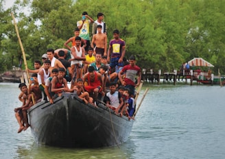 A large group of people, mostly young men, are crowded onto a small boat on a body of water. The boat is moving forward, with some individuals sitting and others standing, while a person in the back takes a photo. Green trees line the distant shore, and a stilted structure is visible in the background.