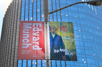 A street banner is displayed against a backdrop of a modern glass building. The banner features two parts: the left side is a red section with white text promoting an Edvard Munch exhibit. The right side shows a colorful painting of a man in a blue coat, with text referencing SF MOMA. The whole scene is lit by daylight, highlighting the urban setting.