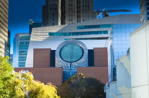 A modern architectural facade features a large circular design with striped patterns, flanked by geometric building elements in shades of brown and blue. The background includes several high-rise buildings with reflective glass windows, and there is a construction crane visible. Trees with green and yellow foliage are present in the foreground.
