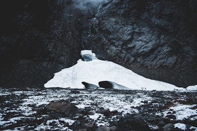 Snow-covered glacier nestled in a rugged mountain landscape, with two visible openings towards the base. Small figures can be seen near the openings, highlighting the vastness of the icy terrain. Jagged rocks and a dark, imposing cliff surround the glacier, creating a dramatic and powerful scene.
