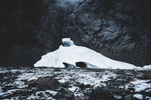 Snow-covered glacier nestled in a rugged mountain landscape, with two visible openings towards the base. Small figures can be seen near the openings, highlighting the vastness of the icy terrain. Jagged rocks and a dark, imposing cliff surround the glacier, creating a dramatic and powerful scene.