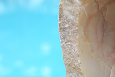 A close-up view of a textured, rough stone surface with a smooth, polished side, displaying various shades of beige and cream. The background is a blurred light blue, possibly resembling water or sky.