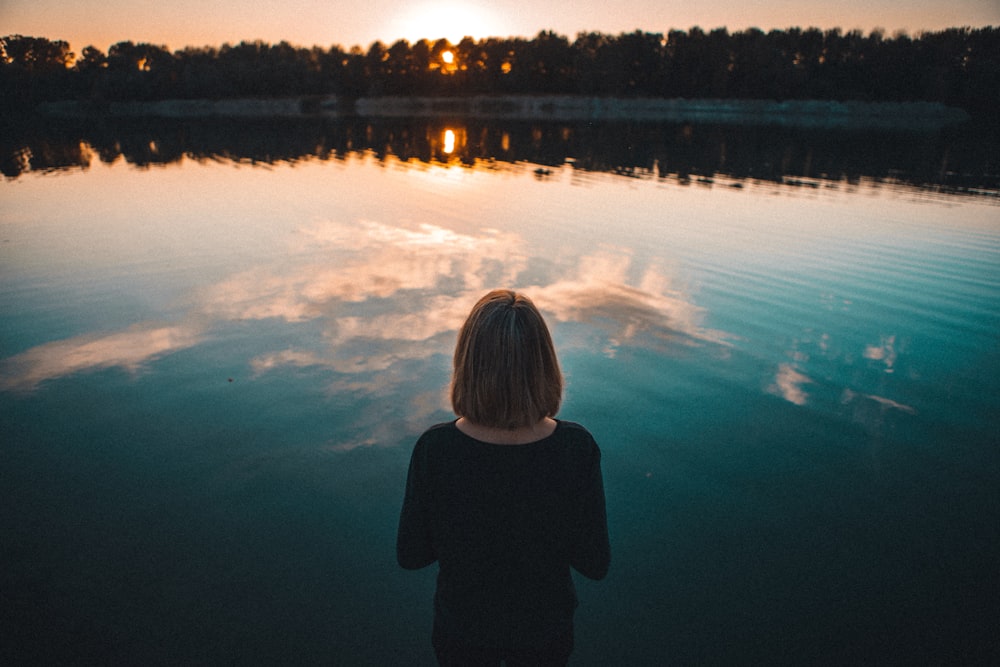 Mujer con vestido negro de pie frente al cuerpo de agua durante la puesta del sol