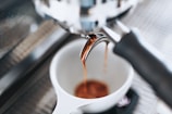 Espresso flows from a portafilter spout into a white cup, highlighting the rich, warm tones of freshly brewed coffee. The setting appears to be a coffee machine, with blurred elements in the background, emphasizing the focus on the coffee-making process.