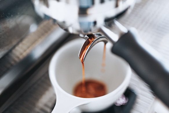 Espresso flows from a portafilter spout into a white cup, highlighting the rich, warm tones of freshly brewed coffee. The setting appears to be a coffee machine, with blurred elements in the background, emphasizing the focus on the coffee-making process.
