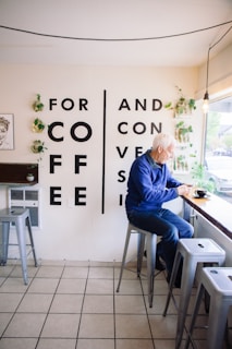 A man sits on a stool at a bar-style counter, facing a window, with a cup of coffee in front of him. The wall is adorned with large bold letters spelling out 'FOR COFFEE AND CONVERSATION'. The setting includes several high stools and potted plants on the wall for decoration.