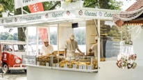 A street food stall selling various baked goods, including pretzels and other pastries. Two workers, wearing white uniforms and caps, are seen behind the glass counter displaying the items. To the left, a classic red and white retro van is parked nearby. The stall signage is decorated with images of pretzels and the word 'Brezel'. In the foreground, heart-shaped gingerbread cookies are hanging, possibly for sale. Trees and a lake can be seen in the background.