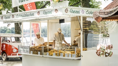 A street food stall selling various baked goods, including pretzels and other pastries. Two workers, wearing white uniforms and caps, are seen behind the glass counter displaying the items. To the left, a classic red and white retro van is parked nearby. The stall signage is decorated with images of pretzels and the word 'Brezel'. In the foreground, heart-shaped gingerbread cookies are hanging, possibly for sale. Trees and a lake can be seen in the background.