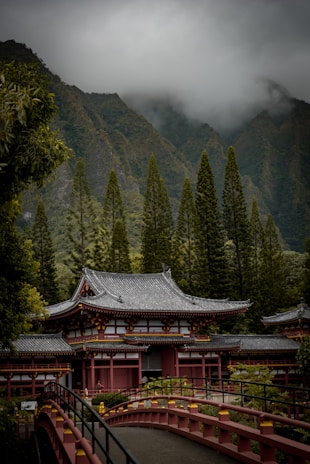 A traditional Japanese-style building is set against a backdrop of lush green mountains and towering trees, with a layer of clouds hanging low. A red arched bridge leads to the entrance of the structure, adding to the serene and tranquil environment.