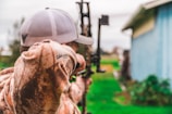 A person wearing a camouflage jacket and a cap is aiming a compound bow outdoors. The background is slightly blurred, showing a green grassy area and part of a blue building.