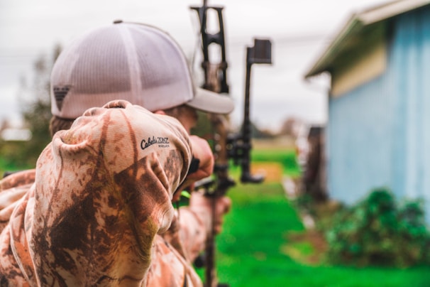 A person wearing a camouflage jacket and a cap is aiming a compound bow outdoors. The background is slightly blurred, showing a green grassy area and part of a blue building.