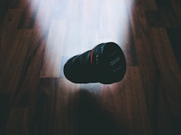A camera lens is placed on a wooden surface, with light casting shadows around it. The lens appears to be in the center of the image, with the brand name visible. The lighting creates a subtle highlight on the wood grain.