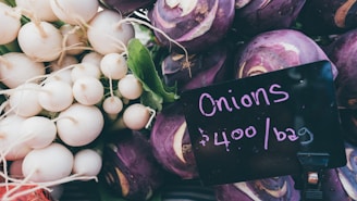 A collection of purple and white vegetables arranged in a market setting. A sign notes 'Onions $400/bag' with white radishes and kohlrabi visible.