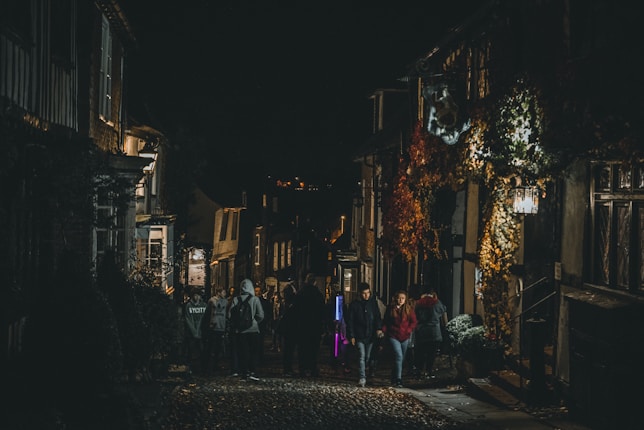 A cobblestone street is dimly lit at night, with groups of people walking along. The street is flanked by old buildings, some covered in autumn foliage. Soft light emanates from windows and street lamps, casting a warm glow.