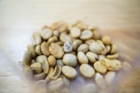 A close-up view of a cluster of unroasted coffee beans against a light wooden background. The beans vary slightly in shape and color, predominantly beige, with one bean appearing slightly darker and more textured than the others.