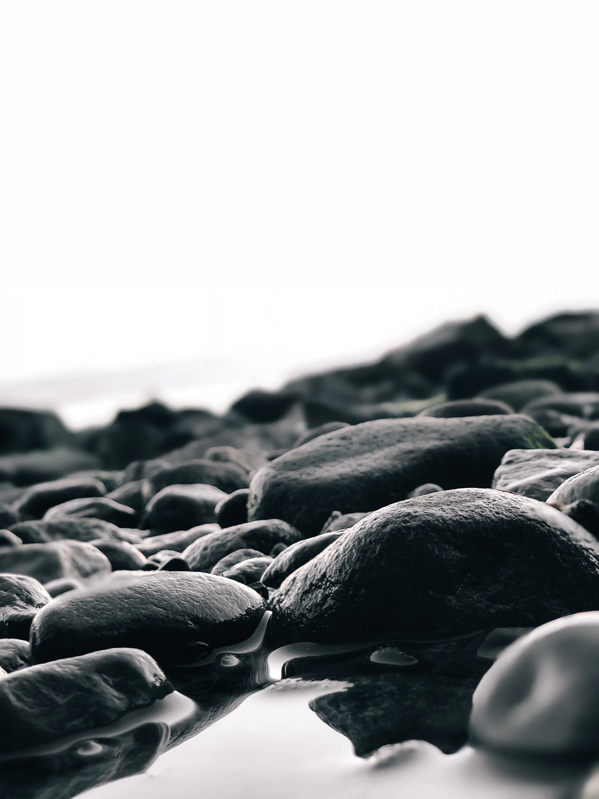 A close-up shot of crystal-clear ocean water swirling around smooth, dark stones on a minimalist beach.