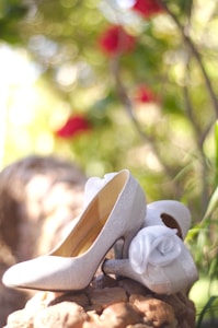 A pair of elegant, glittery high-heeled shoes is positioned outdoors on a textured natural surface. The shoes have decorative white roses attached to them and are illuminated by soft, natural light. In the background, there is a lush bokeh effect with hints of red and green, indicating foliage and possibly flowers.