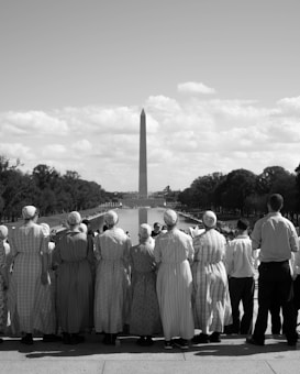 A group of people dressed in traditional, modest clothing stand facing a large reflecting pool, with the Washington Monument prominently visible in the background under a partly cloudy sky.