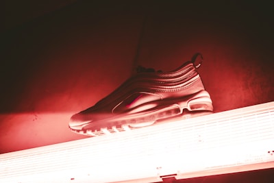 A sneaker placed on top of a fluorescent light, illuminated with a red and pink hue. The sneaker has a sleek design, featuring an air sole and a textured upper.