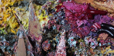 Thick, textured layers of colorful paint are mixed together, creating an abstract and heavily textured appearance. Various hues of pink, red, yellow, purple, blue, and brown form an intricate blend, with some paint strokes showcasing a glossy finish while others appear more matte and rough.