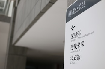 A sign in a modern building displays text in both English and Chinese. It provides directions to the Acquisition & Cataloguing Department, Compact Stacks, and Archives. The setting appears to be a university or a similar institutional environment with a sleek, minimalistic design.