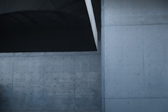 A modern architectural setting with large concrete walls featuring a smooth, minimalistic design. The surfaces are marked with evenly spaced circular indentations, contributing to a sleek, industrial look. The overall structure has a geometric and symmetrical aspect with subtle shadows.