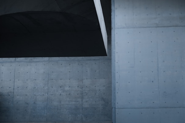 A modern architectural setting with large concrete walls featuring a smooth, minimalistic design. The surfaces are marked with evenly spaced circular indentations, contributing to a sleek, industrial look. The overall structure has a geometric and symmetrical aspect with subtle shadows.