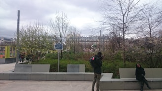 An outdoor urban setting with a person taking photographs and another sitting on a bench. Leafless trees and dense shrubbery occupy the foreground, while residential buildings form the backdrop. The sky is overcast, adding a muted light to the scene.