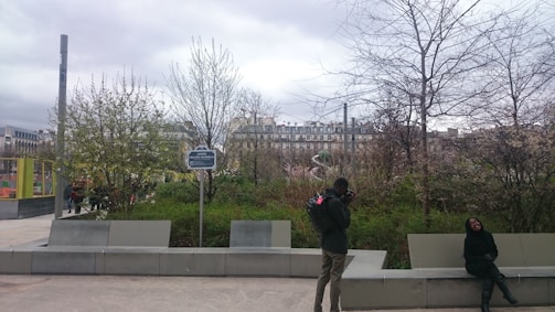 An outdoor urban setting with a person taking photographs and another sitting on a bench. Leafless trees and dense shrubbery occupy the foreground, while residential buildings form the backdrop. The sky is overcast, adding a muted light to the scene.