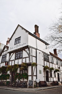 A historic, half-timbered building with a sign reading 'Duke of Wellington' at the top. The structure has exposed wooden beams and white plaster walls. Hanging flower baskets add color to the exterior. Several picnic tables and a menu board are visible outside on the sidewalk. The setting feels quaint and traditional.