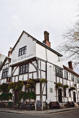 A historic, half-timbered building with a sign reading 'Duke of Wellington' at the top. The structure has exposed wooden beams and white plaster walls. Hanging flower baskets add color to the exterior. Several picnic tables and a menu board are visible outside on the sidewalk. The setting feels quaint and traditional.