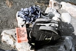 A pebbled shoreline with a black and gray backpack, a checkered shirt, and a book titled 'The Subtle Art of Not Giving a F*ck' by Mark Manson on top of a large rock.