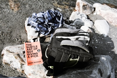A pebbled shoreline with a black and gray backpack, a checkered shirt, and a book titled 'The Subtle Art of Not Giving a F*ck' by Mark Manson on top of a large rock.