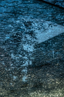 A close-up view of shattered glass with a bluish tint, reflecting light and creating patterns of cracks and fragments. The surface appears rough and uneven, with some areas catching the light more than others.