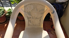 A plastic chair with a floral design on its backrest is placed on a tiled floor. There are plants in pots nearby, and some clothing and a pair of sandals are visible in the background.