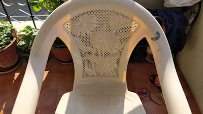 A plastic chair with a floral design on its backrest is placed on a tiled floor. There are plants in pots nearby, and some clothing and a pair of sandals are visible in the background.