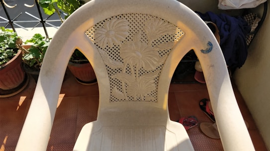 A plastic chair with a floral design on its backrest is placed on a tiled floor. There are plants in pots nearby, and some clothing and a pair of sandals are visible in the background.